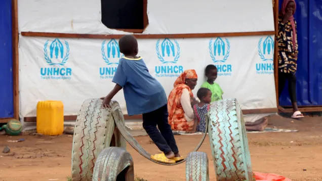 أطفال لاجئون سودانيون وأمهم أمام خيمة بها شعار UNHCR.jpg